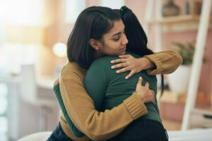 Picture of two young South Asian women hugging in a well lit room. They both have long dark hair. One woman is wearing a green top and has her hair in a ponytail, and the other is wearing dark yellow. The woman in green has her face hidden form the camera.